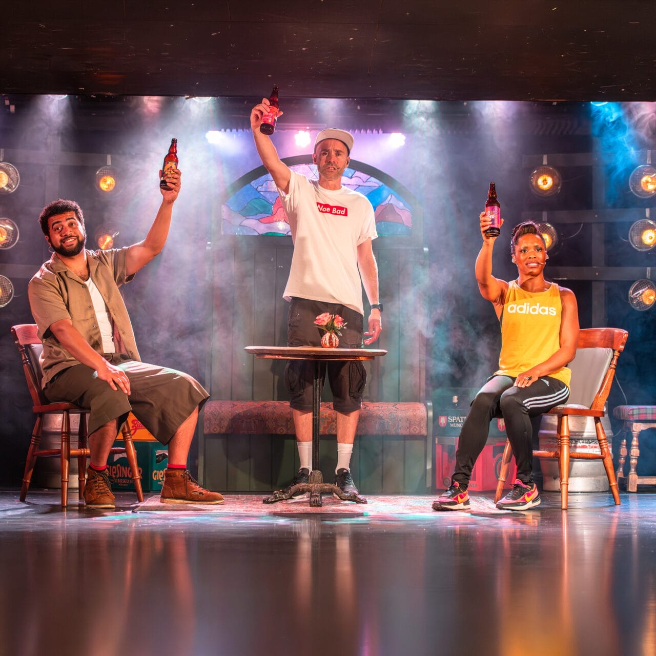 Three people sit and stand around a small table on a stage, each holding up a bottle. Stage lights shine behind them, and there is theatrical smoke in the background. The mood appears celebratory.