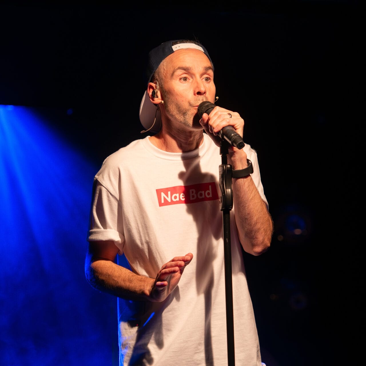 A man wearing a white "Nae Bad" T-shirt and a backwards cap sings into a microphone on stage with blue stage lighting in the background.
