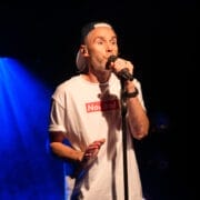A man wearing a white "Nae Bad" T-shirt and a backwards cap sings into a microphone on stage with blue stage lighting in the background.