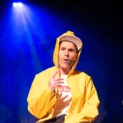 A person wearing a yellow raincoat and a white baseball cap sits under stage lighting, with one hand raised and mouth slightly open as if speaking or singing. The background is dark with blue and purple lights.