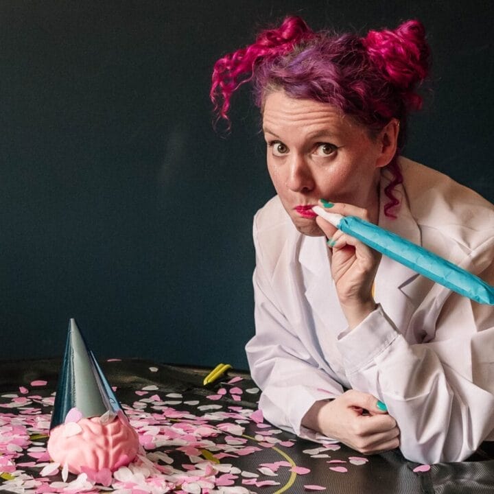 A person with pink and purple hair in a lab coat, blowing a party horn beside a brain model and party hat on a confetti-covered table.