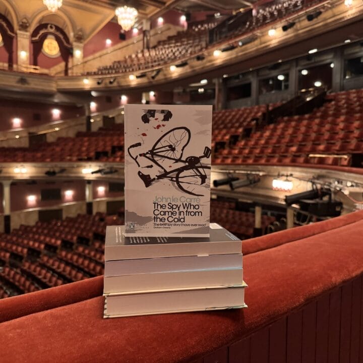 A stack of books, with "The Spy Who Came in from the Cold" by John le Carré on top, rests on a balcony railing inside a large, empty theatre.