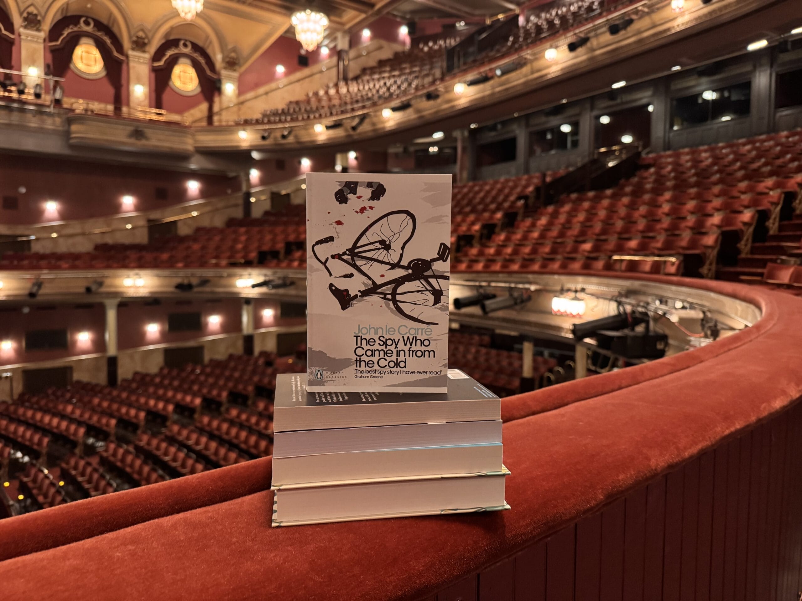 A stack of books, with "The Spy Who Came in from the Cold" by John le Carré on top, rests on a balcony railing inside a large, empty theatre.