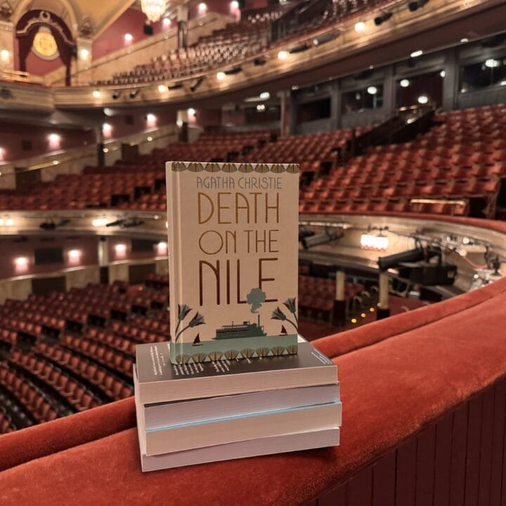 A copy of "Death on the Nile" by Agatha Christie rests on a stack of books atop a red balcony railing inside an empty theatre, ready for the Theatre Book Club.