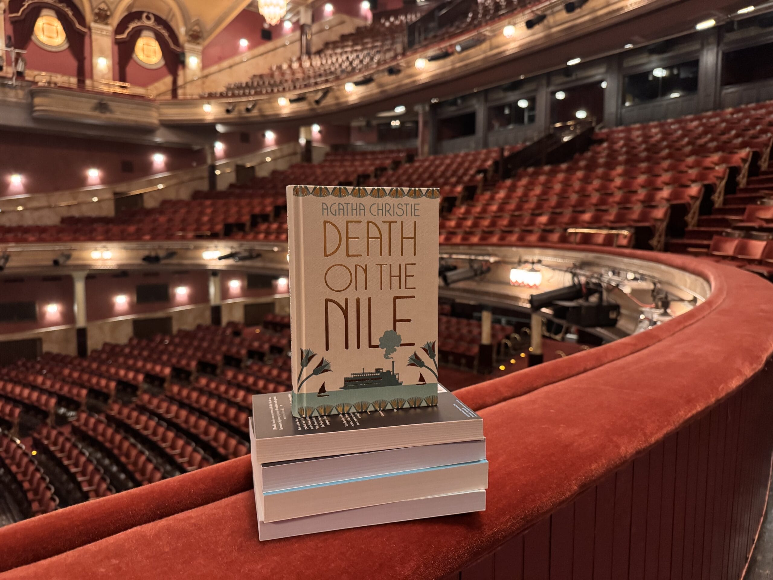 A copy of "Death on the Nile" by Agatha Christie rests on a stack of books atop a red balcony railing inside an empty theatre, ready for the Theatre Book Club.