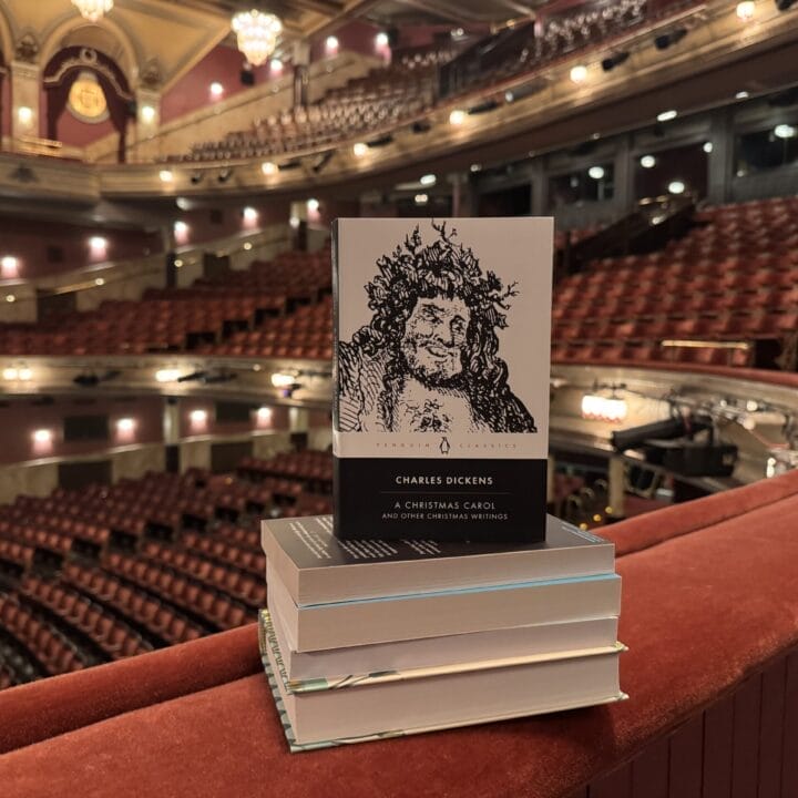 A stack of books sits on a red balcony railing inside an empty theatre, with "A Christmas Carol" by Charles Dickens visible on top.
