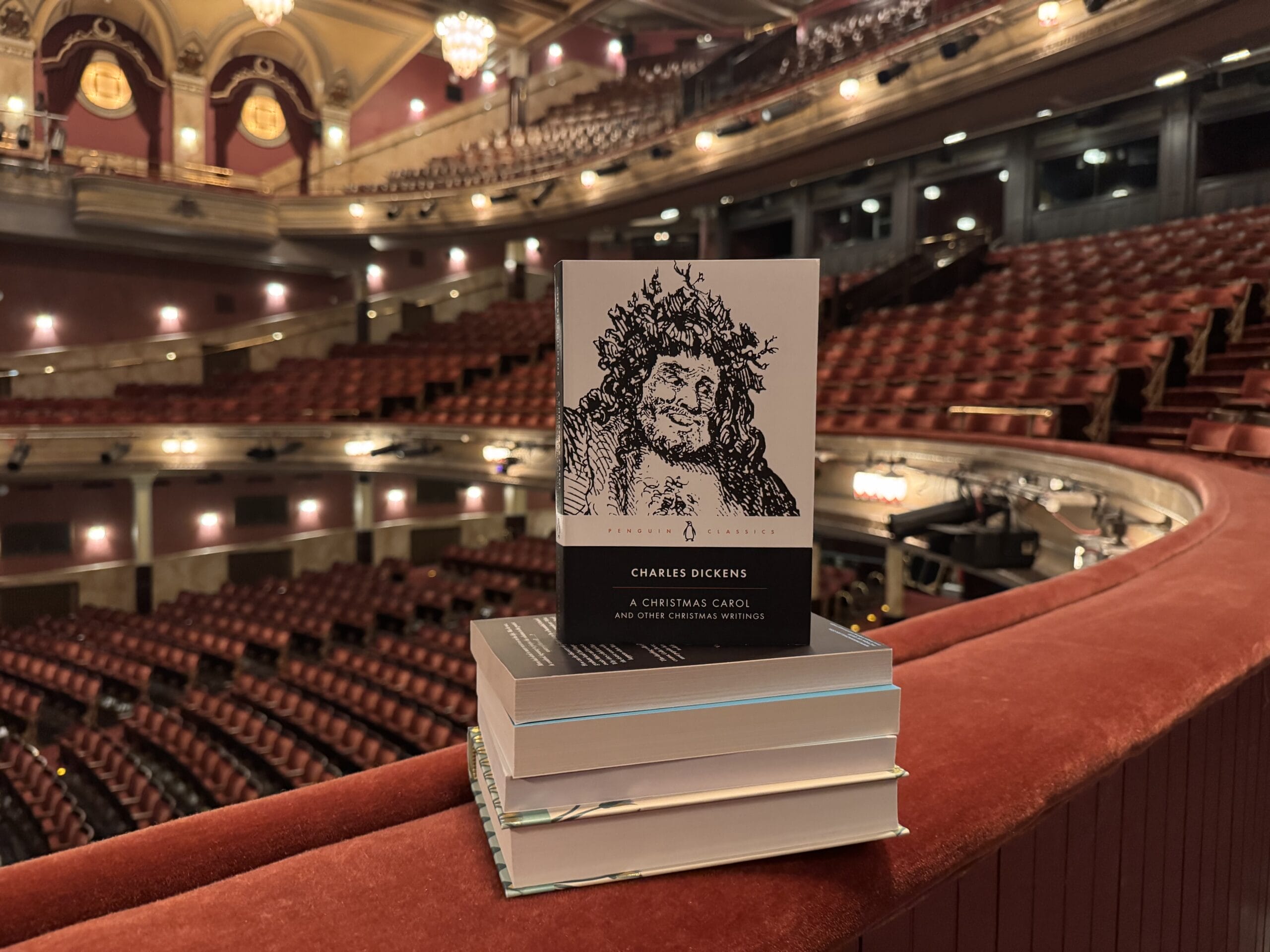 A stack of books sits on a red balcony railing inside an empty theatre, with "A Christmas Carol" by Charles Dickens visible on top.