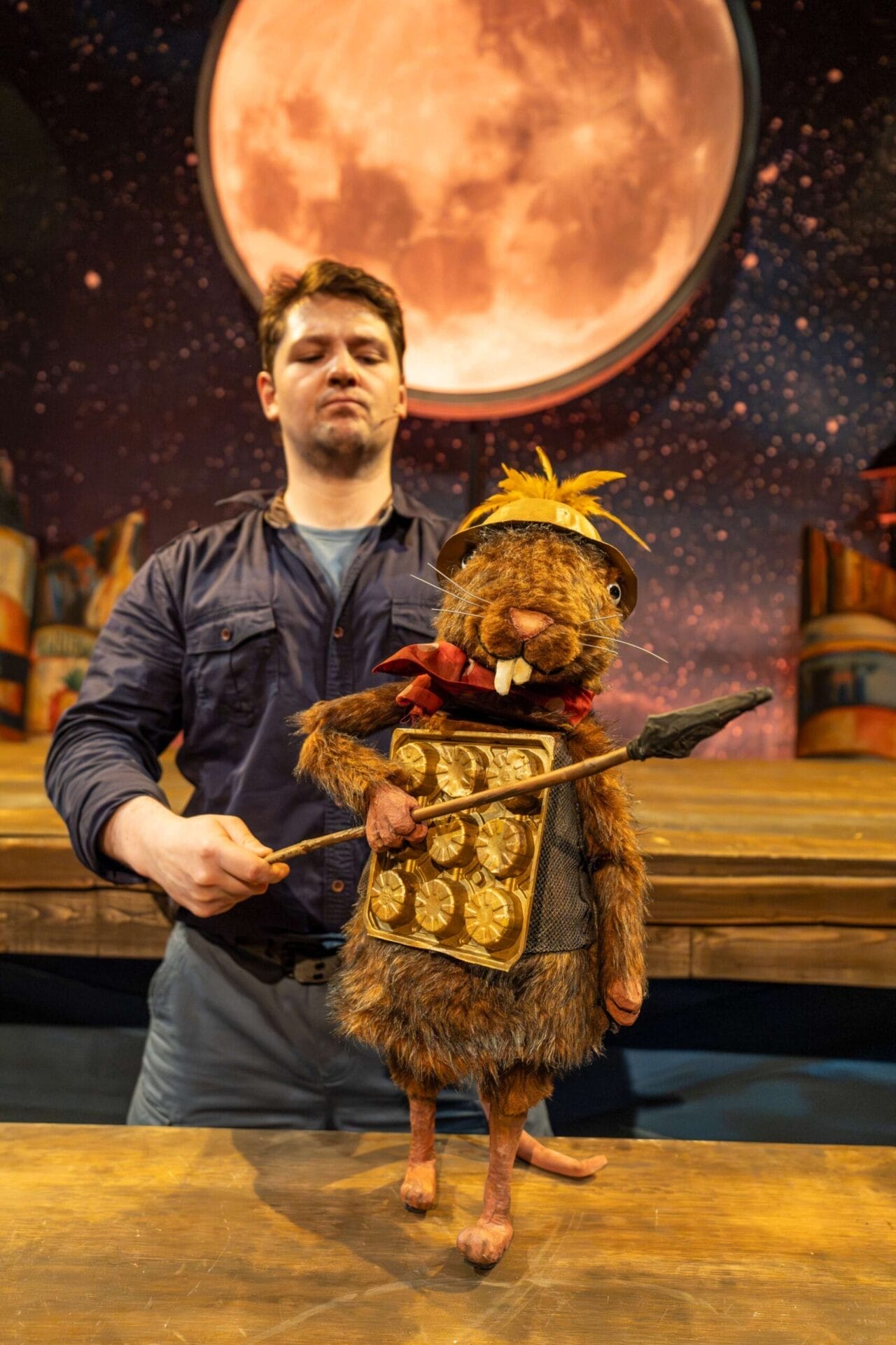 A man operates a beaver puppet dressed as a warrior, holding a shield and spear, in front of a stage set with a large, illuminated moon and starry sky