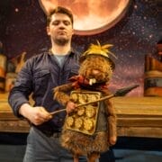 A man operates a beaver puppet dressed as a warrior, holding a shield and spear, in front of a stage set with a large, illuminated moon and starry sky