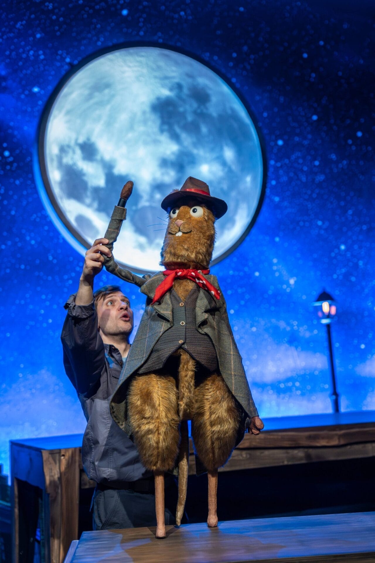 A puppeteer operates a large, well-dressed animal puppet resembling Toto the Ninja Cat, complete with a red scarf and hat, in front of a moonlit night sky backdrop during a stage performance.