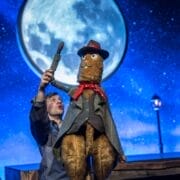 A puppeteer operates a large, well-dressed animal puppet resembling Toto the Ninja Cat, complete with a red scarf and hat, in front of a moonlit night sky backdrop during a stage performance.