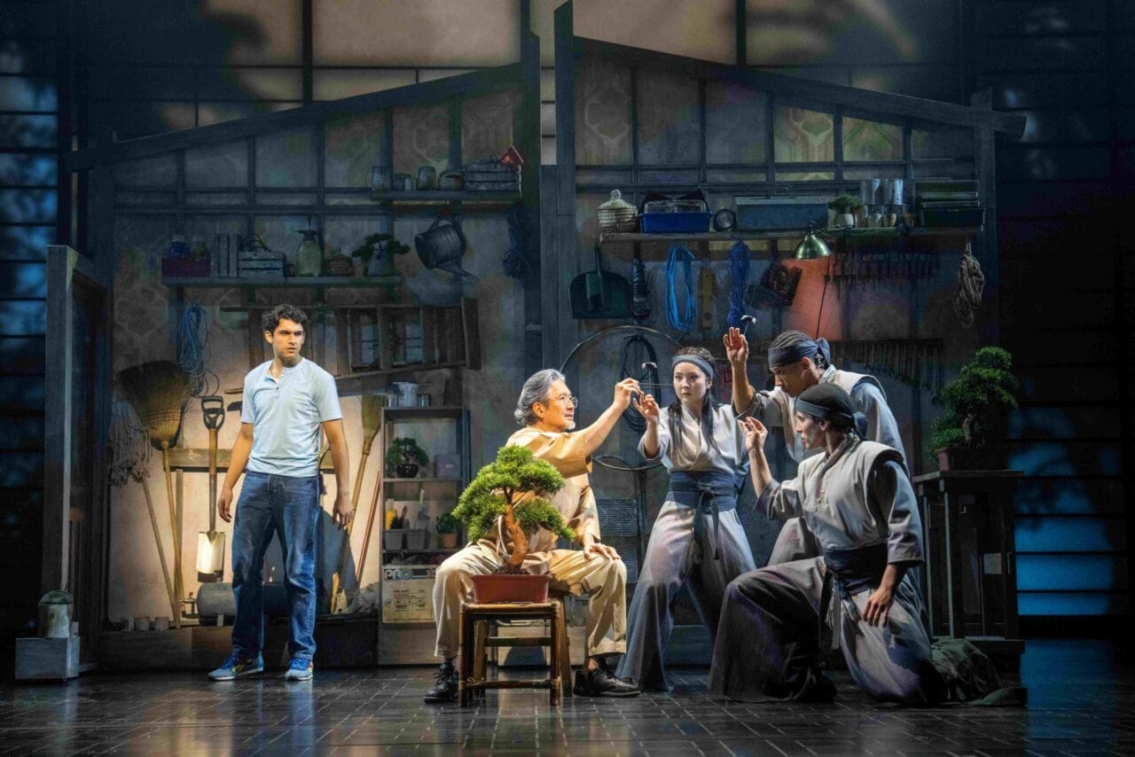 Five actors perform on stage in a workshop set with tools and plants. One, seated at a table with a bonsai tree, evokes memories of The Karate Kid whilst the others engage in a dramatic scene reminiscent of The Musical’s energy.