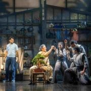 Five actors perform on stage in a workshop set with tools and plants. One, seated at a table with a bonsai tree, evokes memories of The Karate Kid whilst the others engage in a dramatic scene reminiscent of The Musical’s energy.