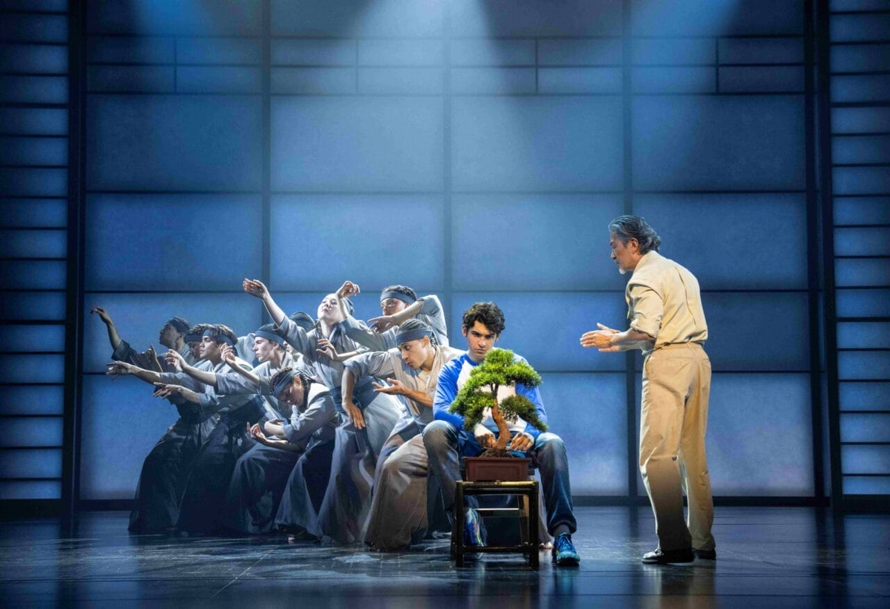 A theatrical stage scene inspired by The Karate Kid shows a group dramatically reaching towards a young man in a blue shirt with a potted plant, whilst another man gestures beside him. Blue-toned lighting adds a moody, musical effect.