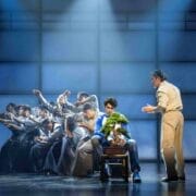 A theatrical stage scene inspired by The Karate Kid shows a group dramatically reaching towards a young man in a blue shirt with a potted plant, whilst another man gestures beside him. Blue-toned lighting adds a moody, musical effect.