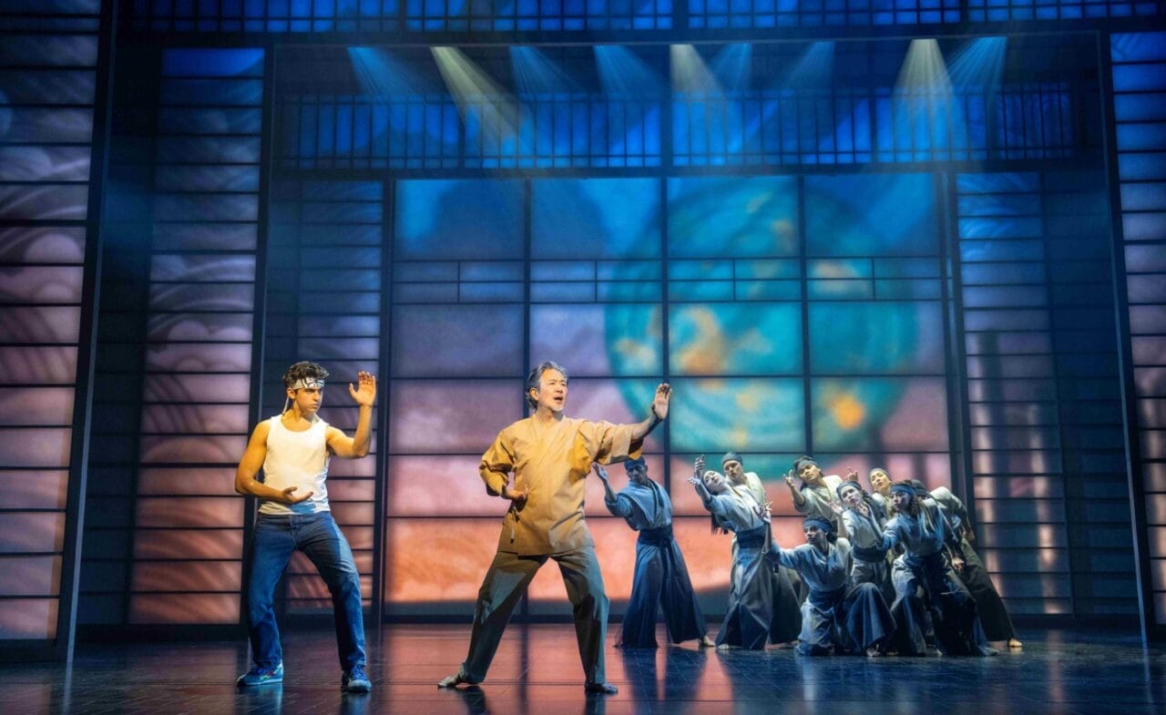 Several performers are on stage, with two people in martial arts poses at the front and a group in coordinated stances behind them, capturing the energetic spirit of The Karate Kid Musical amid dramatic lighting and abstract designs.