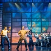 Several performers are on stage, with two people in martial arts poses at the front and a group in coordinated stances behind them, capturing the energetic spirit of The Karate Kid Musical amid dramatic lighting and abstract designs.