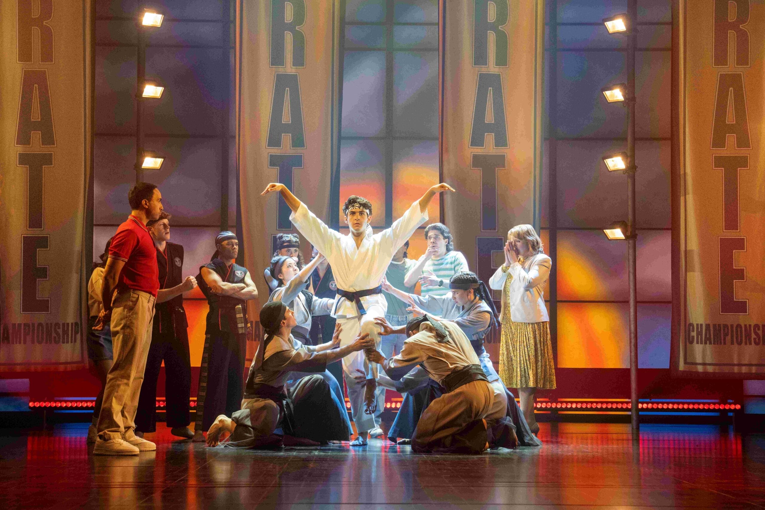 A group of people on stage surround a person in a white karate uniform posing with arms raised, evoking The Karate Kid Musical. Large banners reading "KARATE" and "CHAMPIONSHIPS" hang in the brightly lit background.
