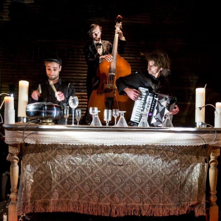 Five musicians perform around a table with candles, playing violin, glass harp, double bass, accordion, and cello, with a patterned cloth draped over the table.