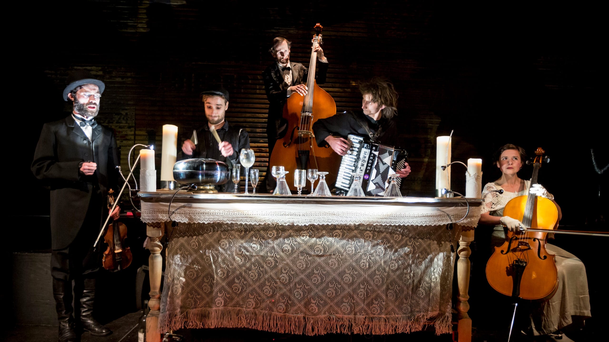 Five musicians perform around a table with candles, playing violin, glass harp, double bass, accordion, and cello, with a patterned cloth draped over the table.