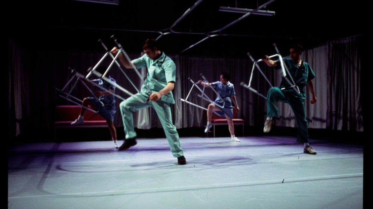 Four people in medical scrubs perform a synchronized dance on stage, each holding and lifting a metal chair, with curtains and overhead lights in the background.