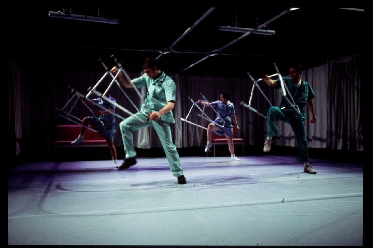Four people in medical scrubs perform a synchronized dance on stage, each holding and lifting a metal chair, with curtains and overhead lights in the background.