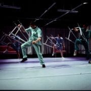 Four people in medical scrubs perform a synchronized dance on stage, each holding and lifting a metal chair, with curtains and overhead lights in the background.