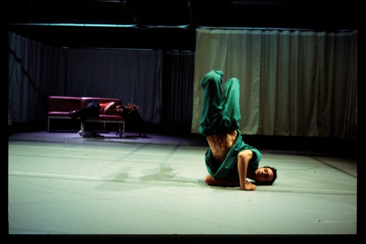 A person in green clothing performs a headstand on a stage, while another person lies on a couch in the background.