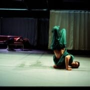 A person in green clothing performs a headstand on a stage, while another person lies on a couch in the background.