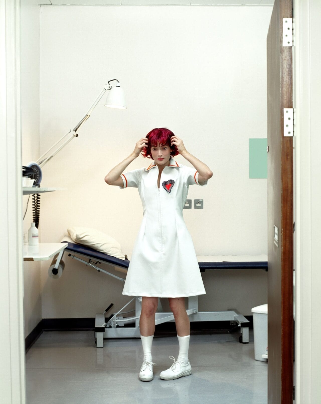 A woman with bright red hair in a white dress with a heart patch stands in a sparse medical examination room, facing the camera and adjusting her hair.