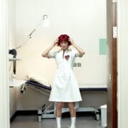 A woman with bright red hair in a white dress with a heart patch stands in a sparse medical examination room, facing the camera and adjusting her hair.