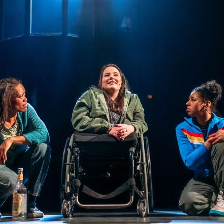 Three women sit on stage under dramatic lighting at King’s Theatre; the woman in the center is in a wheelchair, reflecting theatre accessibility. The others kneel on either side, with a bottle on the floor next to them.