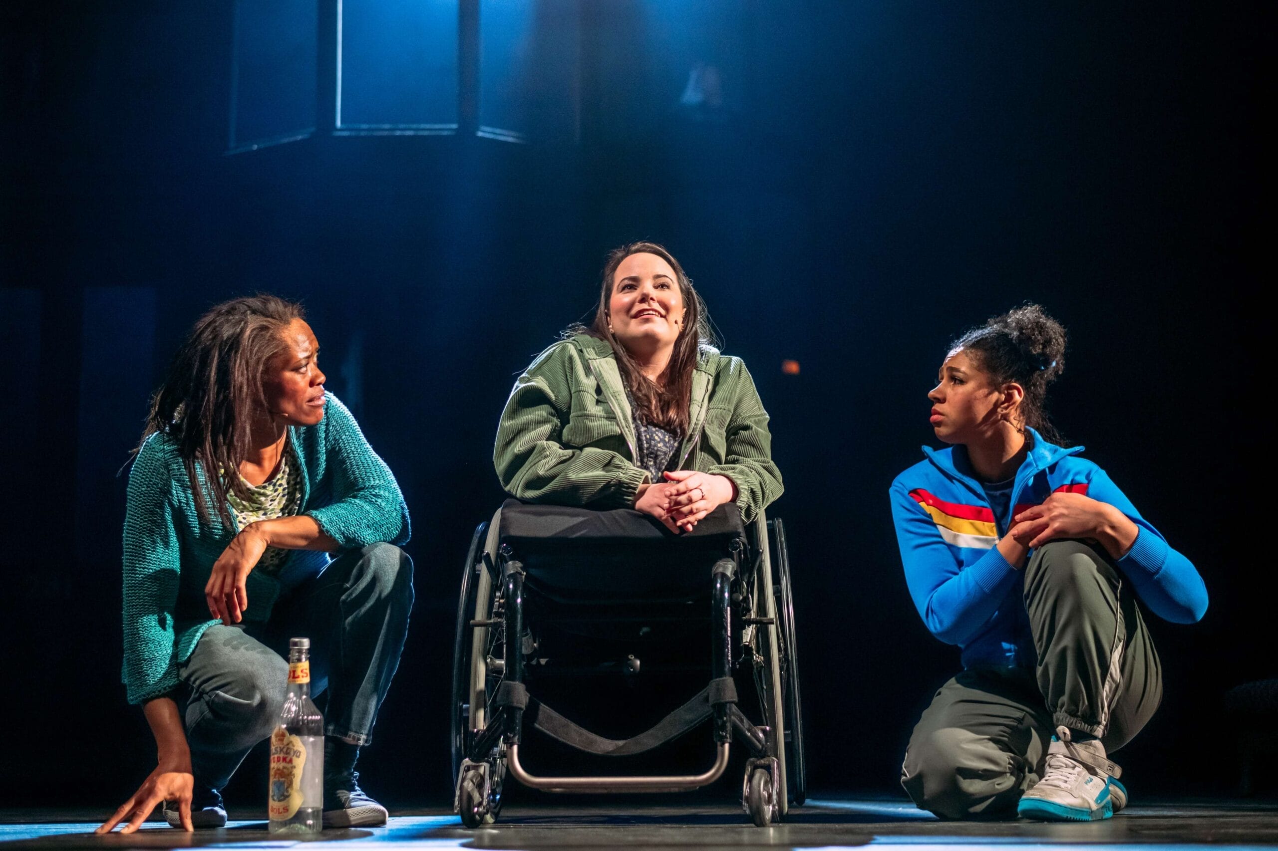 Three women sit on stage under dramatic lighting at King’s Theatre; the woman in the center is in a wheelchair, reflecting theatre accessibility. The others kneel on either side, with a bottle on the floor next to them.
