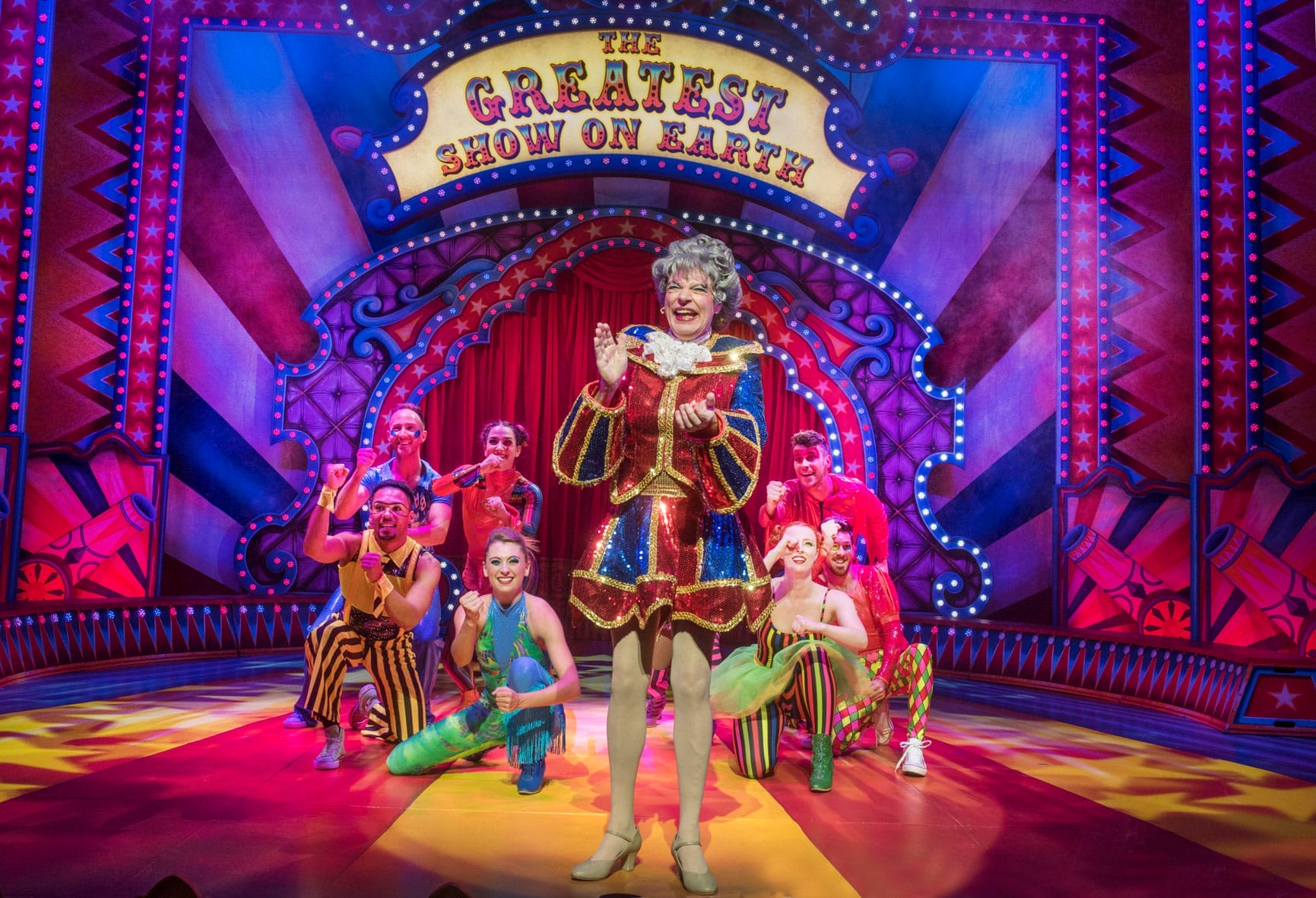A group of performers in colorful circus costumes surround Allan Stewart as Dame Auntie May on stage under a sign reading "The Greatest Show on Earth."
