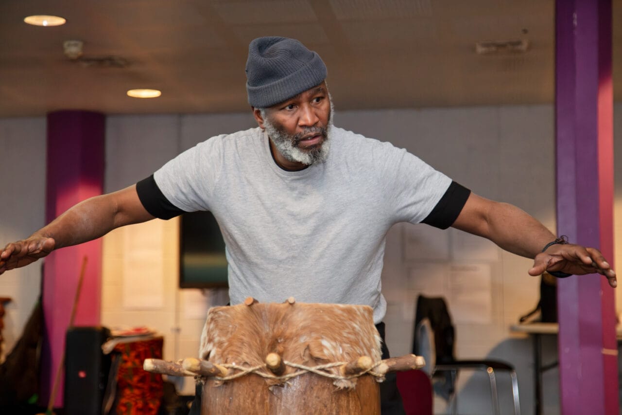 A man wearing a gray beanie and t-shirt stands with arms outstretched behind a large Crown of Blood drum in an indoor setting.