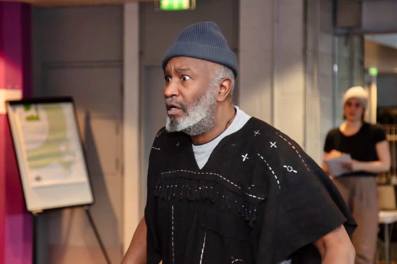 A man with a gray beard wearing a knit hat and a patterned black poncho stands indoors, looking concerned, as if burdened by a Crown of Blood. In the background, a person holds papers.