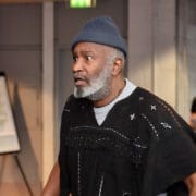 A man with a gray beard wearing a knit hat and a patterned black poncho stands indoors, looking concerned, as if burdened by a Crown of Blood. In the background, a person holds papers.