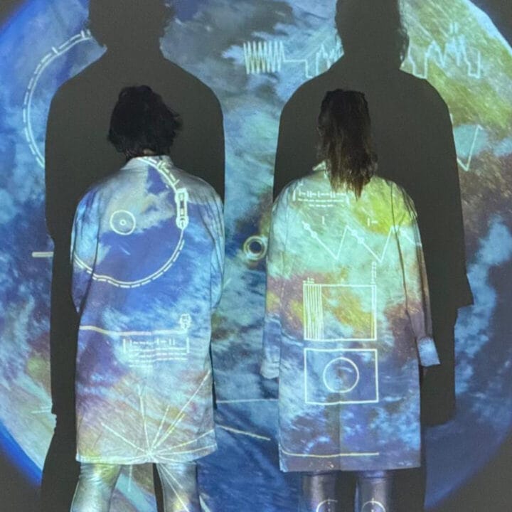 Two people in white coats stand with their backs to the camera, with imagery of Earth and scientific diagrams projected onto them and the wall behind.