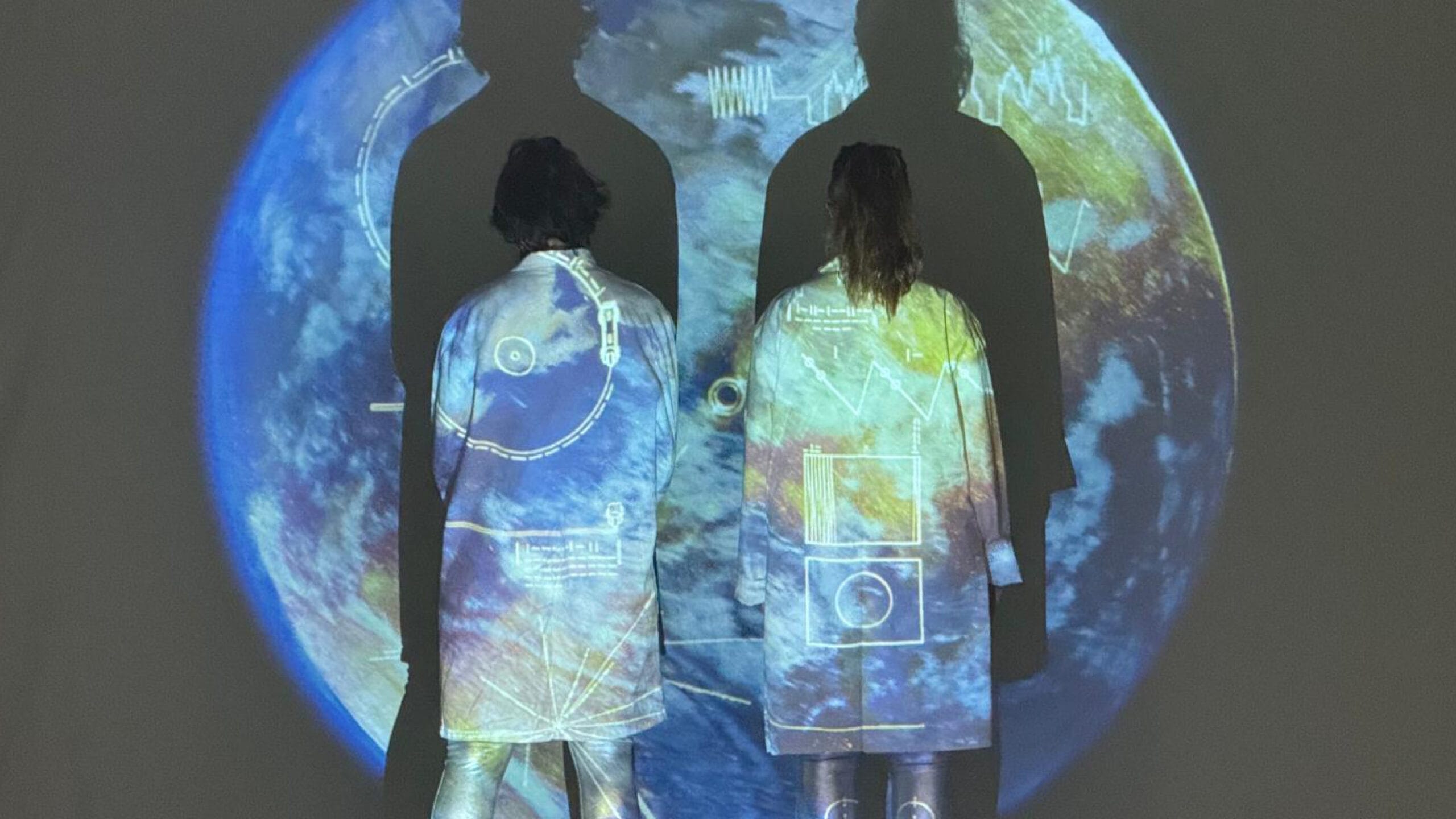Two people in white coats stand with their backs to the camera, with imagery of Earth and scientific diagrams projected onto them and the wall behind.