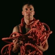 A person stands against a dark background holding a large bundle of thick red rope draped over their arms and shoulders.