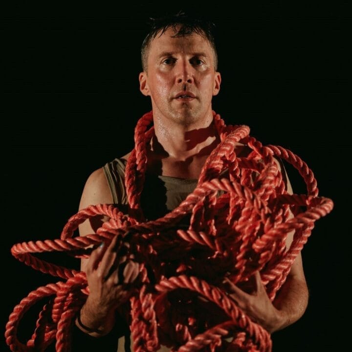A person stands against a dark background holding a large bundle of thick red rope draped over their arms and shoulders.