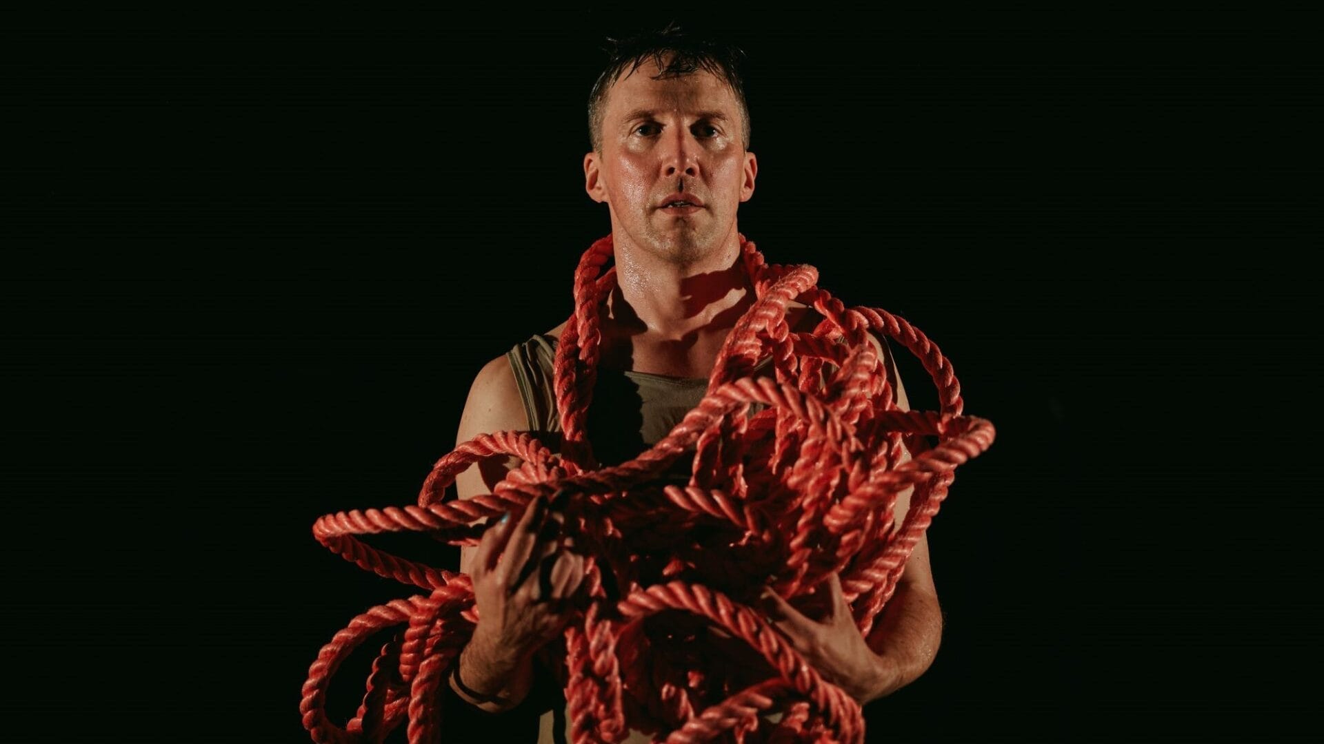 A person stands against a dark background holding a large bundle of thick red rope draped over their arms and shoulders.
