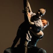A person performs with a human-like puppet, both suspended against a dark background.