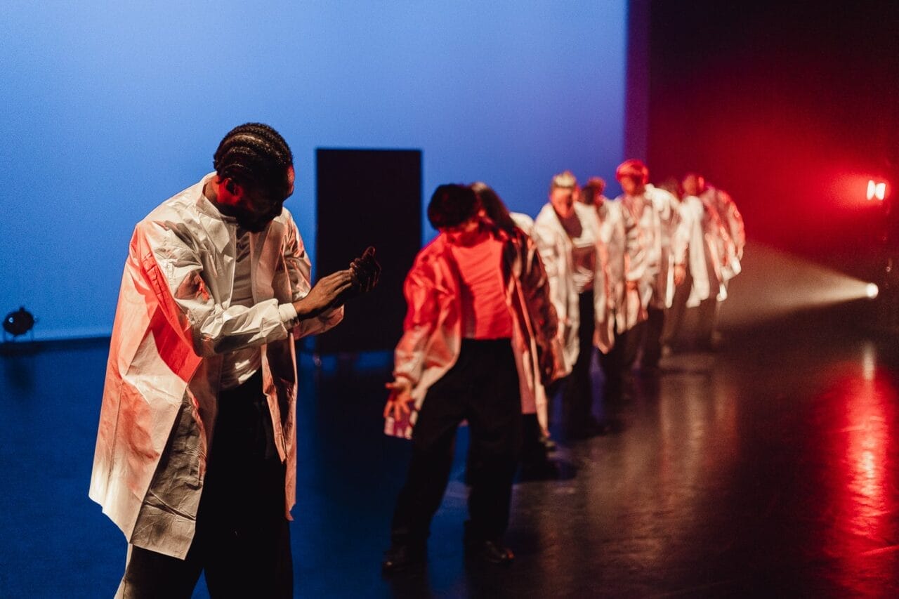 A line of dancers in white jackets perform on a dimly lit stage with blue and red lighting, showcasing Evolution by Three60 World. The front dancer is clapping, whilst others are positioned behind, facing different directions.