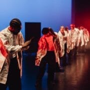 A line of dancers in white jackets perform on a dimly lit stage with blue and red lighting, showcasing Evolution by Three60 World. The front dancer is clapping, whilst others are positioned behind, facing different directions.