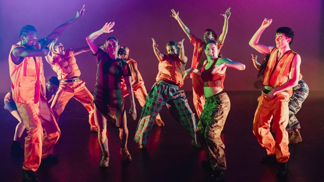 A group of dancers wearing bright orange outfits and patterned clothing perform energetic movements on stage, illuminated by colourful lighting, showcasing the spirit of Evolution powered by Three60 World.