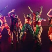 A group of dancers wearing bright orange outfits and patterned clothing perform energetic movements on stage, illuminated by colourful lighting, showcasing the spirit of Evolution powered by Three60 World.