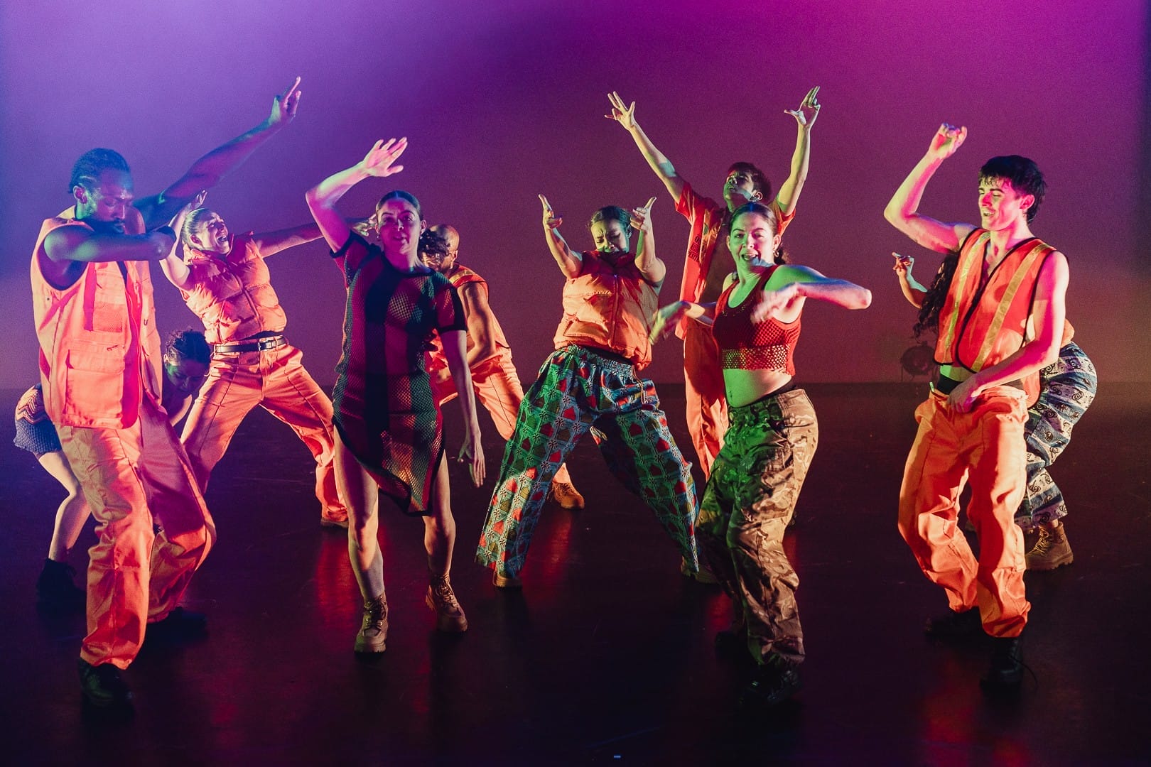 A group of dancers wearing bright orange outfits and patterned clothing perform energetic movements on stage, illuminated by colourful lighting, showcasing the spirit of Evolution powered by Three60 World.