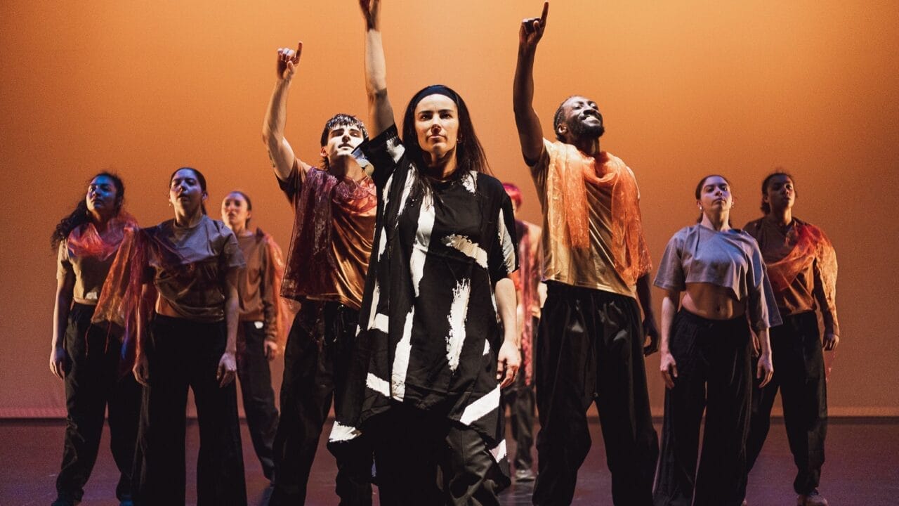 A group of dancers stands on stage under warm orange lighting, embodying the evolution of movement. At the centre, one wears black and white, while others don loose black trousers and sheer orange scarves—many with an arm raised, pointing up. Three60 World energy fills the scene.