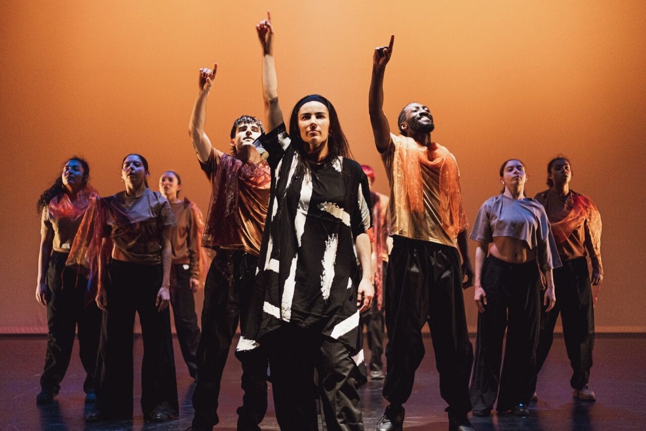 A group of dancers stands on stage under warm orange lighting, embodying the evolution of movement. At the centre, one wears black and white, while others don loose black trousers and sheer orange scarves—many with an arm raised, pointing up. Three60 World energy fills the scene.